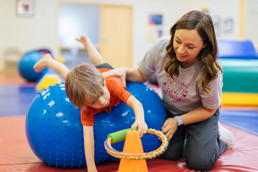 Pediatric therapist working with a young child on core strengthening and coordination using a therapy ball and cones.
